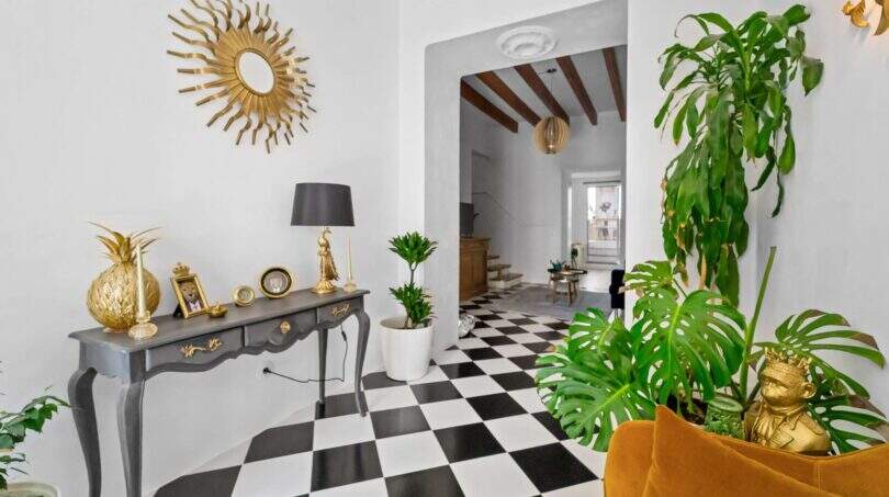 Elegante entrada con piso de tablero de ajedrez, acentos dorados, plantas Monstera y vista al siguiente ambiente