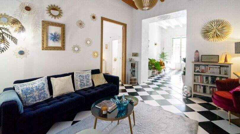 Sala de estar luminosa con piso de tablero de ajedrez, sofá de terciopelo azul, espejos de pared dorados y techo de vigas de madera en casa urbana Felanitx