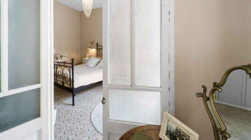 Vista a través de puerta de cristal al dormitorio con cama de hierro forjado, piso de mosaico y lámpara de araña en encantadora casa urbana Felanitx