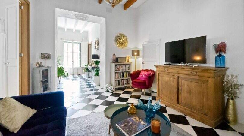 Sala de estar con piso de tablero de ajedrez, techo de vigas de madera, sofá azul, sillón rojo y aparador rústico en casa urbana Felanitx