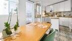 Comedor luminoso con mesa de madera, sillas verdes y cocina abierta con armarios blancos en casa urbana Felanitx