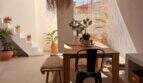 Luminosa zona de comedor con mesa de madera, lámpara de ratán, escalera y plantas en la encantadora casa urbana de Felanitx