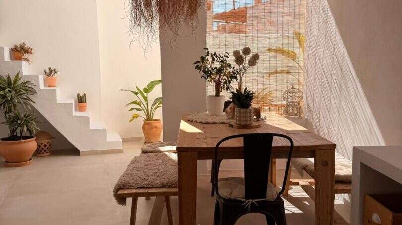 Luminosa zona de comedor con mesa de madera, lámpara de ratán, escalera y plantas en la encantadora casa urbana de Felanitx