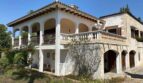 Zweigeschossige mediterrane Villa in Porto Cristo mit Arkaden, Balkon und Natursteinanbau unter blauem Himmel.
