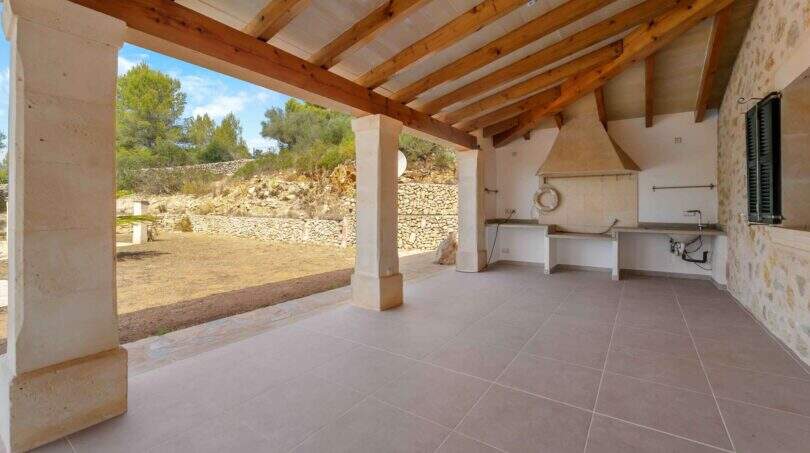 Überdachte Außenküche mit Holzbalkendecke, Natursteinwänden und Gartenblick der Finca in Cala Murada
