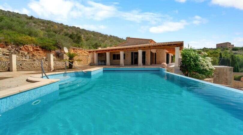 Großer Außenpool der Finca in Cala Murada mit Blick auf das Steinhaus und bewaldete Hügel.