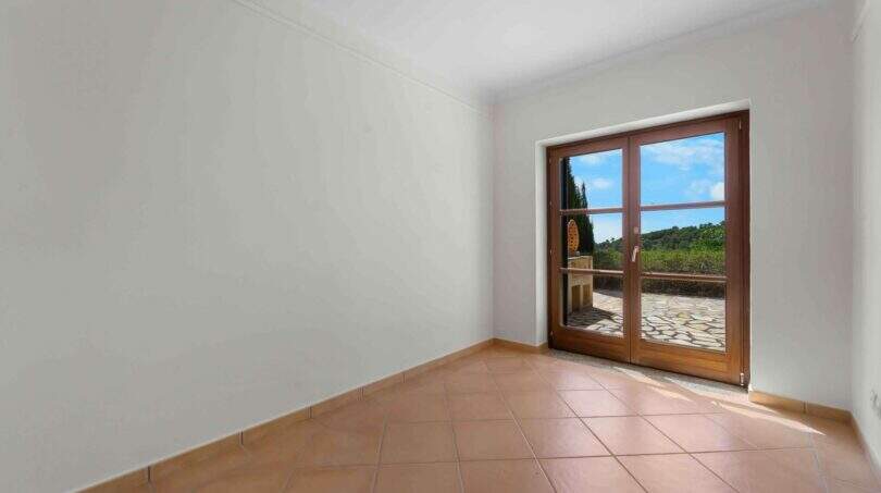 Leeres Zimmer der Finca Cala Murada mit Terrakottaboden und Glastür zum sonnigen Außenbereich mit Grünblick.