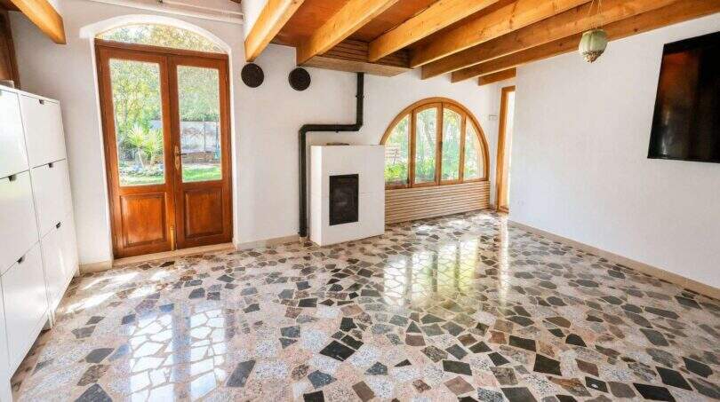 Helles Wohnzimmer der Finca Algaida mit Natursteinboden, Holzbalkendecke, Kamin und Bogenfenstern zum Garten.
