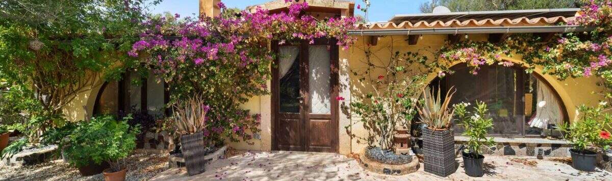 Außenansicht Finca Algaida: mediterrane Villa mit Bougainvillea, Holztür und üppigem Grün unter blauem Himmel.
