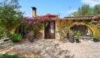 Außenansicht Finca Algaida: mediterrane Villa mit Bougainvillea, Holztür und üppigem Grün unter blauem Himmel.
