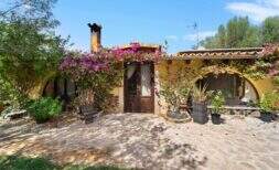 Außenansicht Finca Algaida: mediterrane Villa mit Bougainvillea, Holztür und üppigem Grün unter blauem Himmel.