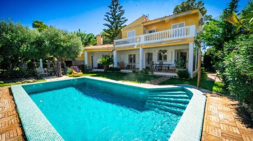 Mallorca-Villa in Llucmajor mit türkisblauem Pool, Holzterrasse, Balkon und üppigem Garten unter blauem Himmel.
