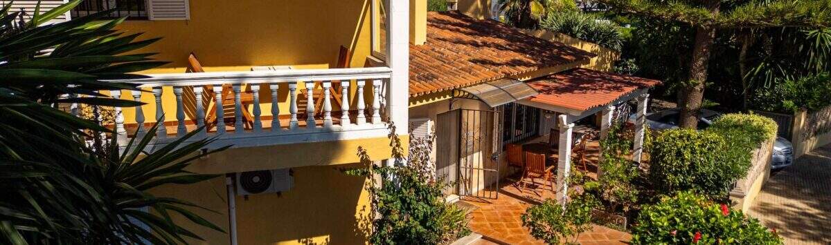 Mediterrane Villa in Llucmajor mit gelbem Fassade, Balkon, Terrakotta-Dach und üppigem Garten mit Palmen.