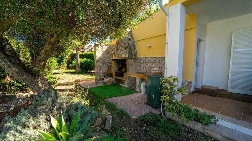 Steinbarbecue im Garten der Mallorca-Villa in Llucmajor, umgeben von üppiger mediterraner Vegetation und gelbem Hausanbau.
