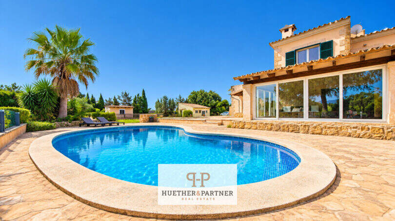 Finca Santa Maria: Pool mit Natursteintetrasse, mediterrane Villa mit Glasfront und Palmen im Sonnenschein