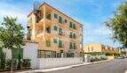 Vista exterior del apartamento esquinero de nueva construcción en Cala Millor, edificio multifamiliar amarillo con persianas verdes y balcones, escalera de caracol.