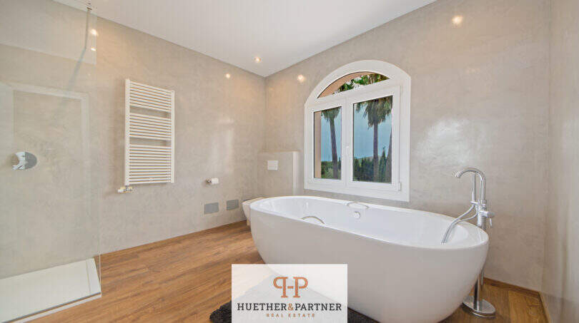 Luxusbad der Finca Santa María: freistehende Badewanne, Glasdusche, Holzboden und Fenster mit Palmenblick.