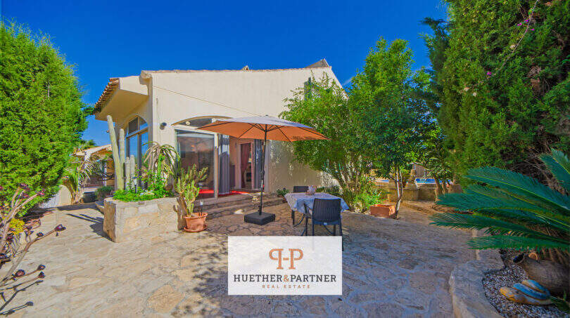 Charmantes Ferienhaus in Cala Murada mit mediterraner Terrasse, Sonnenschirm, Kakteen und üppigem Grün unter blauem Himmel.