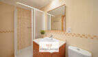 Helles Badezimmer der Erdgeschosswohnung Cala Antena mit Dusche, Waschtisch und beigen Mosaikfliesen.
