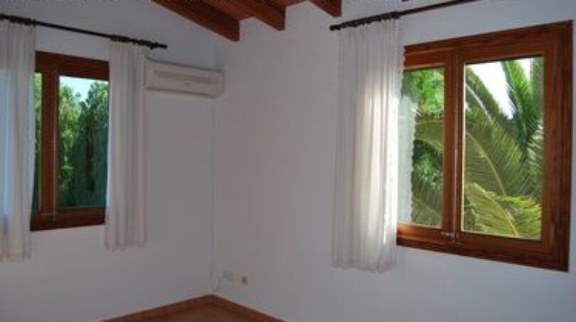Helles Schlafzimmer der Finca Manacor mit Holzfenstern, Klimaanlage und Blick auf tropischen Garten mit Palmen.