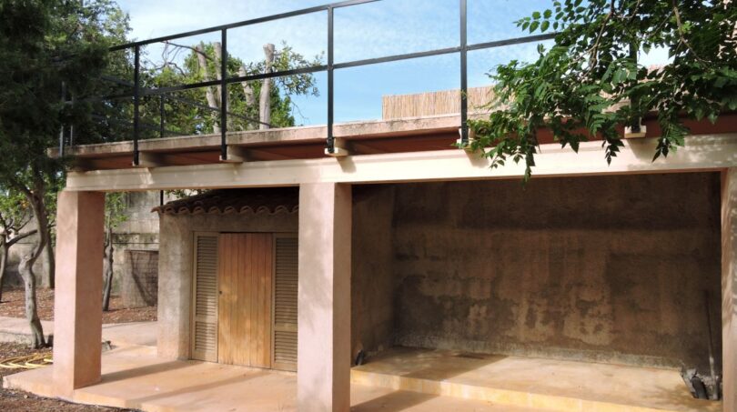 Überdachter Carport mit Dachterrasse und schwarzem Metallgeländer, umgeben von Bäumen der Finca in Manacor