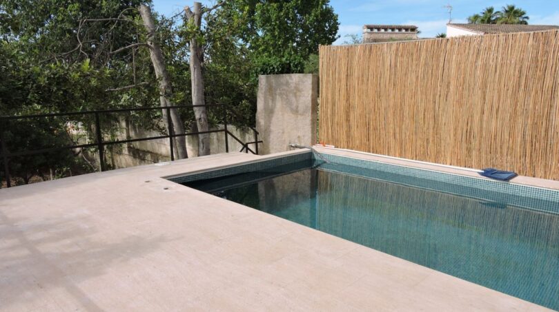 Pool der Finca in Manacor mit hellem Fliesenrand, Schilfmatte als Sichtschutz und üppigem Baumbestand