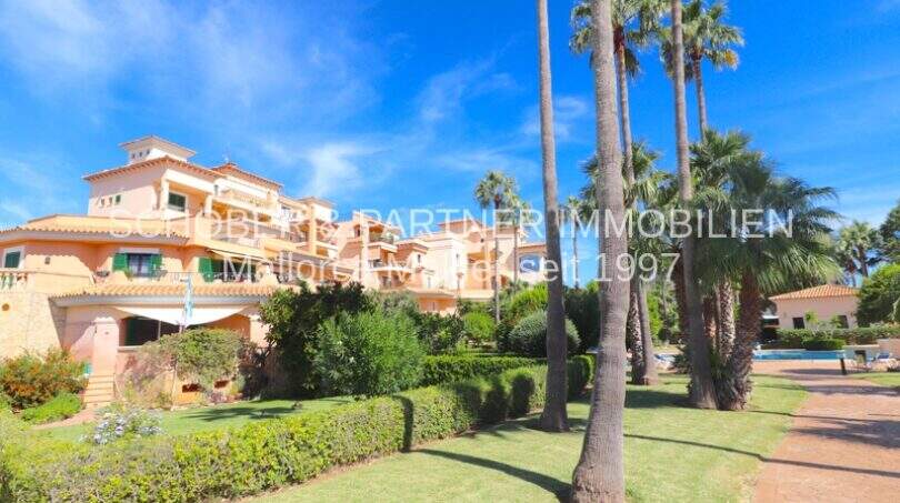 Fachada del complejo residencial Cala Millor: arquitectura mediterránea, jardín cuidado con palmeras y piscina.