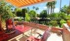 Terraza soleada con mesa de madera, sillas rojas y toldo, vista al exuberante jardín con palmeras en Cala Millor.