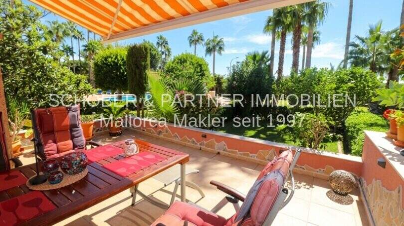 Terraza soleada con mesa de madera, sillas rojas y toldo, vista al exuberante jardín con palmeras en Cala Millor.