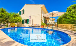 Vista exterior de la casa Cala Murada con piscina ovalada, sombrilla y jardín mediterráneo bajo un cielo azul