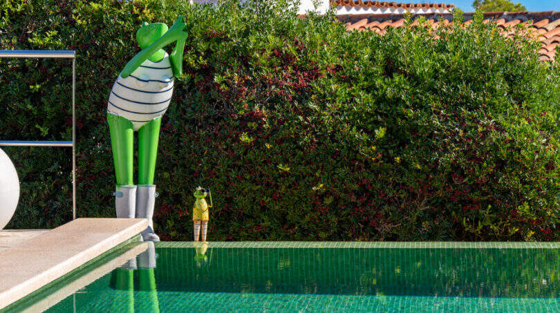 Infinity pool with mirrored water surface and decorative frog sculptures, surrounded by lush hedge at Villa Cala Murada