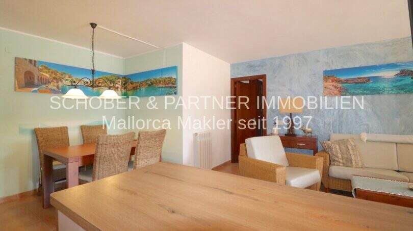 Helles Wohn- und Esszimmer der Poolwohnung in Sa Coma mit Holztisch, Rattanstühlen und mediterraner Wanddekoration.