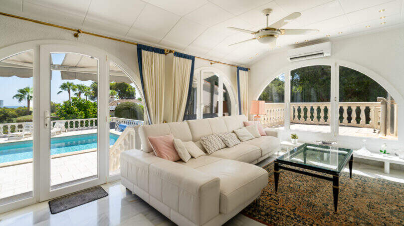 Helles Wohnzimmer der Poolvilla in Cala Murada mit Bogenfenstern, weißem Ecksofa und Blick auf den Pool.