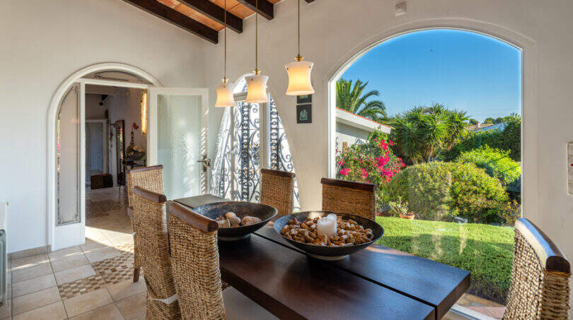 Covered terrace with dining table, wicker chairs and view into the Mediterranean garden of the villa in Cala Murada