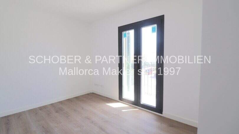 Dormitorio claro y vacío con suelo de madera y puertas-ventana negras en apartamento esquinero de nueva construcción en Cala Millor