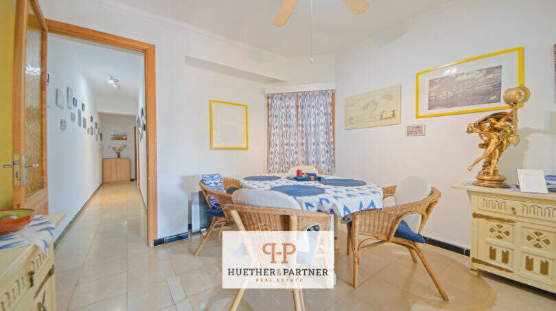Luminoso comedor con sillas de ratán, mantel azul, ventilador de techo y cuadros en la pared en apartamento de playa Porto Cristo