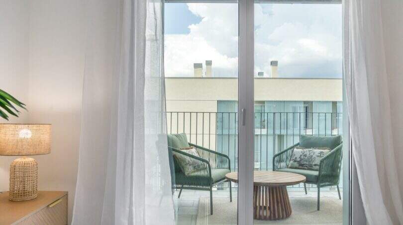 Área de estar luminosa con puerta de cristal al balcón amueblado, tumbonas verdes de descanso y mesa de madera en Palma de Mallorca