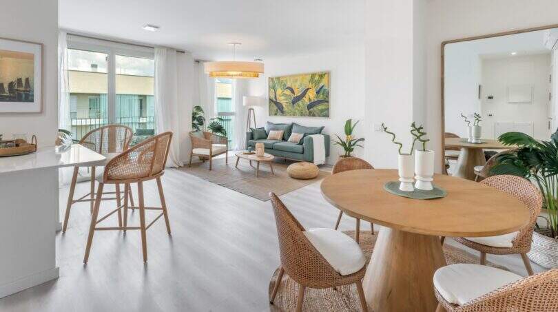 Área de estar-comedor luminosa con muebles de ratán, suelo de madera y grandes ventanas en apartamento exclusivo en Palma