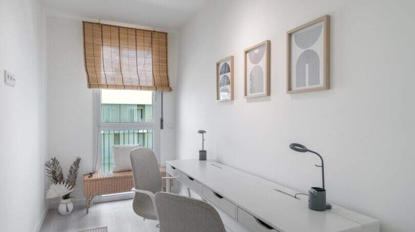 Despacho luminoso con escritorio blanco, sillas grises, estor de bambú e imágenes de pared modernas en apartamento en Palma.