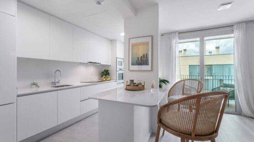 Cocina blanca moderna con isla de cocción, sillas de ratán y acceso a la terraza en apartamento exclusivo, Palma de Mallorca.