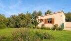 Außenansicht Finca Mallorca: Beige Bungalow mit Holzfensterläden, üppigem Garten, Blumen und Obstbäumen unter blauem Himmel.
