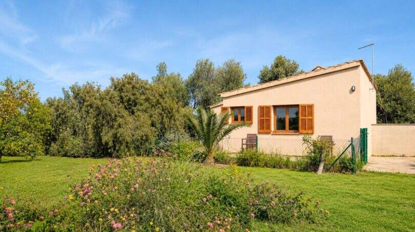 Außenansicht Finca Mallorca: Beige Bungalow mit Holzfensterläden, üppigem Garten, Blumen und Obstbäumen unter blauem Himmel.