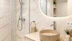 Moderno baño de ático con paredes de mármol, lavabo redondo sobre encimera, espejo iluminado y ducha de cristal.