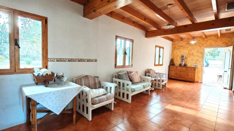Salón luminoso de la finca Inca con techo de vigas de madera, muebles de estar blancos y suelo de terracota