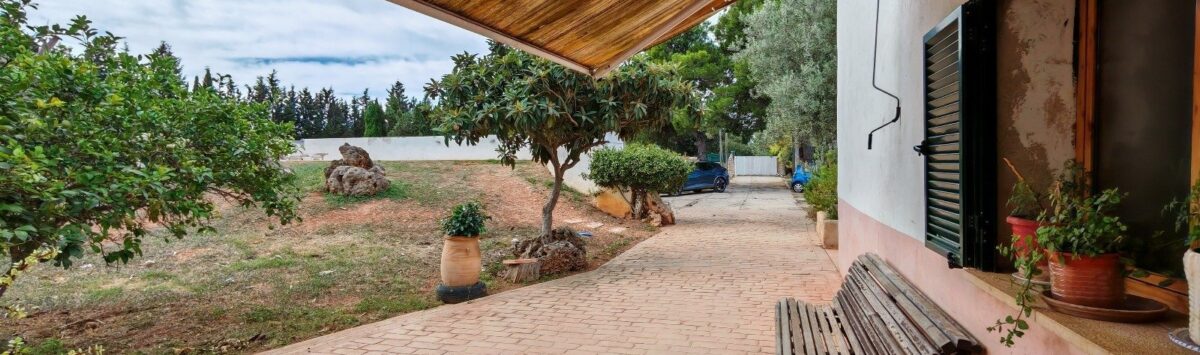 Terraza cubierta de la finca cerca de Inca con banco de madera, adoquines de barro y exuberante jardín con árboles frutales