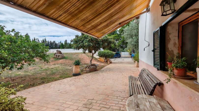 Terraza cubierta de la finca cerca de Inca con banco de madera, adoquines de barro y exuberante jardín con árboles frutales