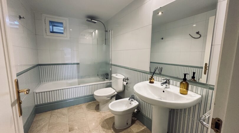 Luminoso baño con bañera, bidé, WC y lavabo, paredes con azulejos blanco-verdes, ventana
