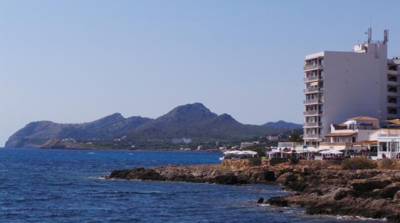 Küstenblick in Cala Ratjada: Felsige Meeresküste mit Hotelgebäude und Bergen im Hintergrund, Mallorca.