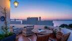 Terraza soleada de villa en Alcanada con muebles de mimbre, macetas y vista panorámica al mar al atardecer.