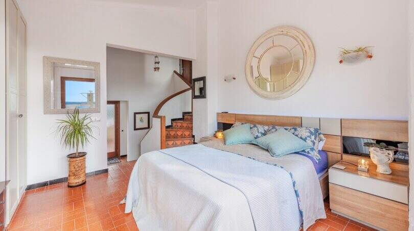 Dormitorio luminoso de villa mediterránea con suelo de terracota, cama doble, escaleras y espejo con vista al mar